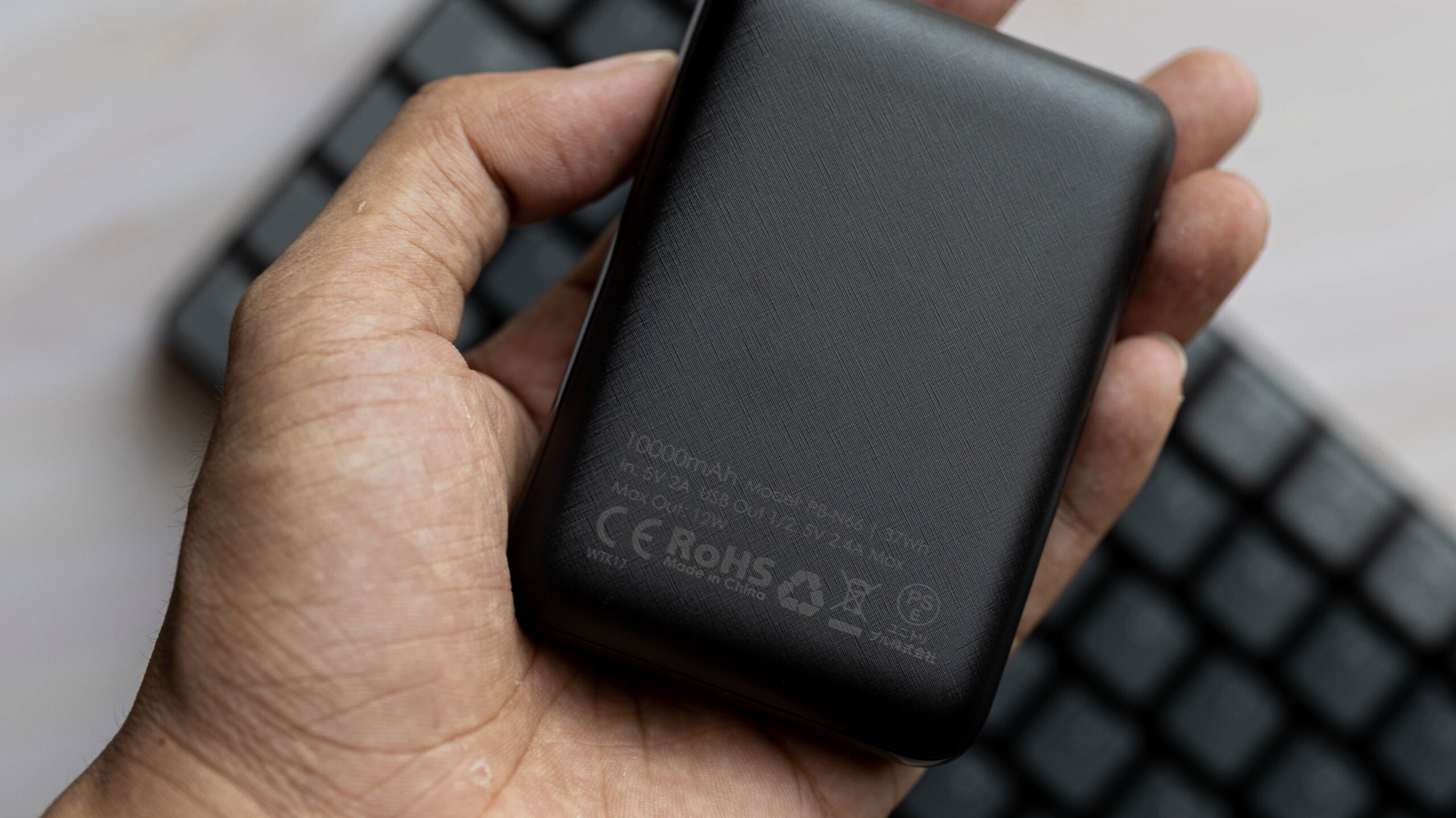 A close-up shot of a hand holding a black power bank with a keyboard in the background.
