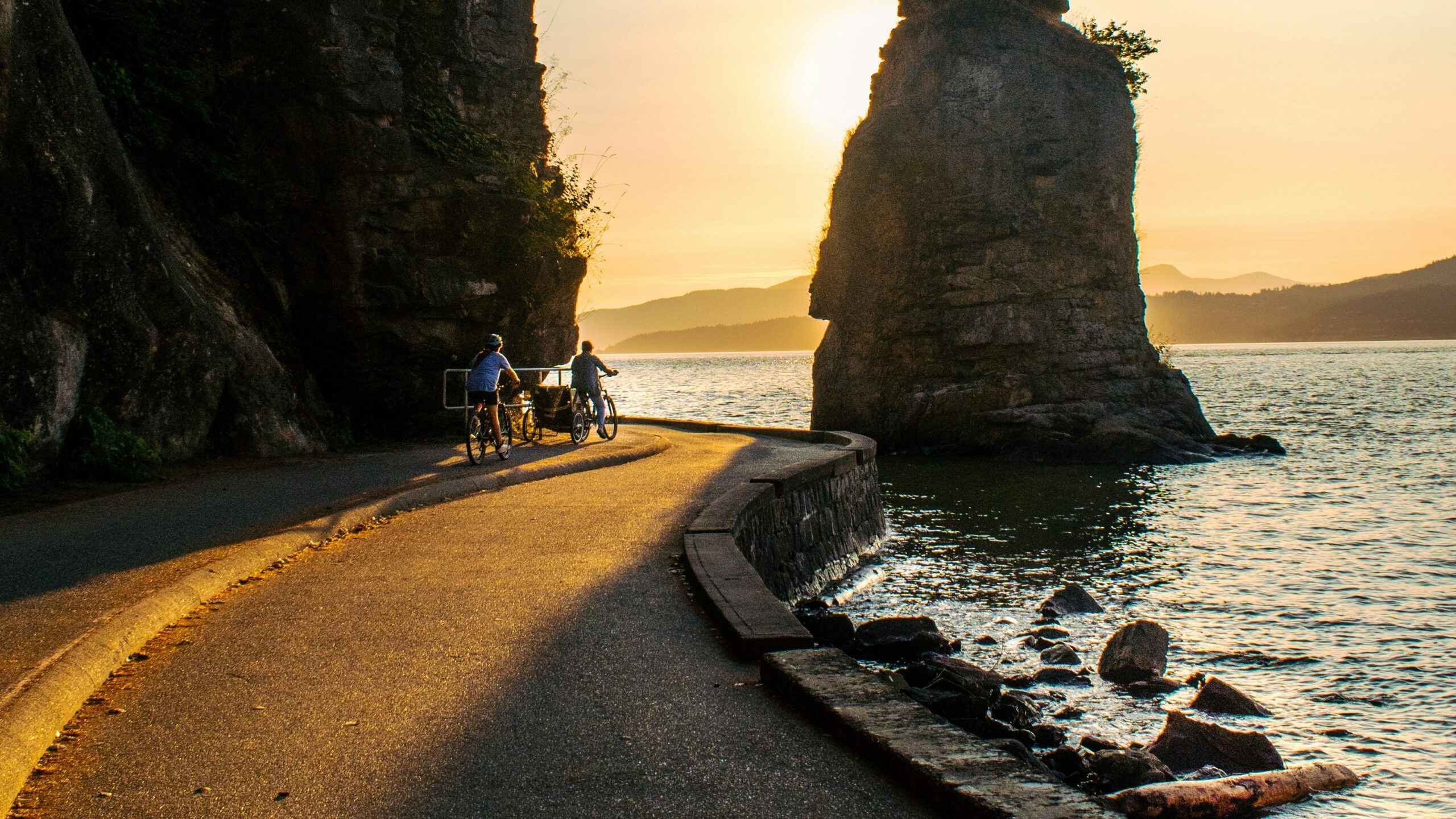 a couple of people riding bikes down a road next to a body of water a couple of people riding bikes down a road next to a body of water