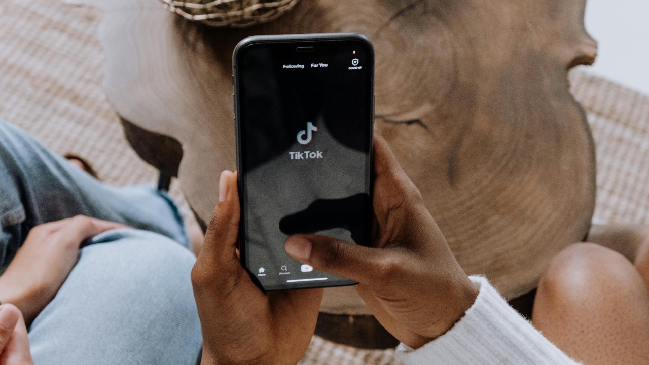 Close-up of hands holding a smartphone displaying the TikTok app interface on a wooden surface. cottonbro studio/Pexels